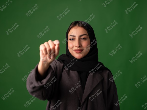 A portrait of a young woman wearing a black hijab and a dark coat against a green background. The woman is smiling softly and appears poised. Her hand is raised in a gentle gesture, adding a sense of engagement.