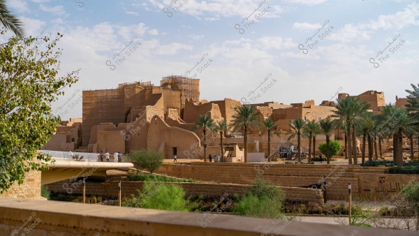 The image shows a historical site in Saudi Arabia. The old mud-brick buildings feature traditional Najdi architecture. Palm trees are visible in the foreground with a blue sky, and scaffolding in the background indicates restoration work.