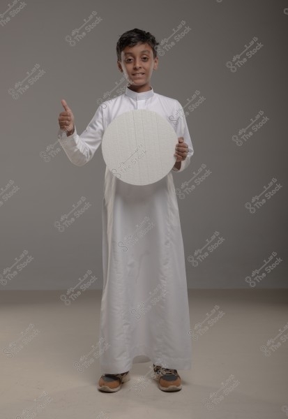An image of a boy wearing a traditional white Arabic thobe, standing in a photography studio with a gray background. The boy is holding a white circular object in his left hand and is giving a thumbs-up with his right hand while smiling. He is wearing athletic shoes.