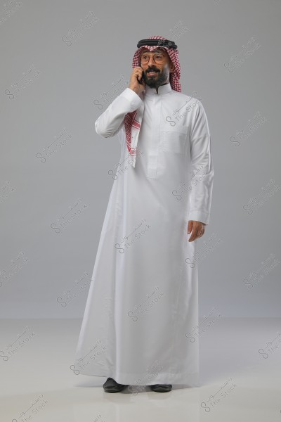 A portrait of a man wearing a white thobe and a traditional headscarf with an agal, talking on a mobile phone. The background is neutral, and the man is slightly smiling, wearing glasses. The scene might be set in Saudi Arabia or another Gulf country.