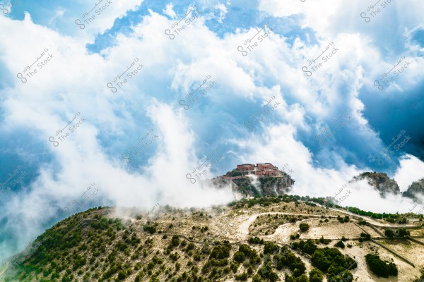 A natural landscape showing thick clouds over mountains covered with greenery. In the background, buildings are visible atop a mountain, surrounded by the dense clouds. The horizon is filled with blue sky and some white clouds, reflecting the beauty of nature at high altitudes.