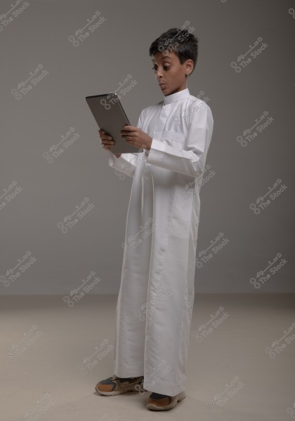 A boy wearing traditional white attire is holding a tablet. He is standing in a side pose in an indoor setting with a neutral gray background, appearing surprised or intrigued by something on the screen.