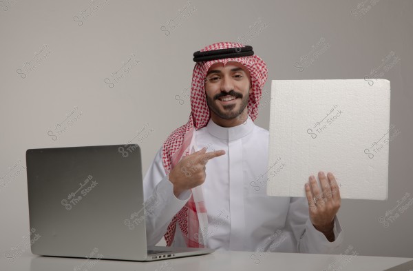 An image of a man sitting in front of a laptop. He is wearing a white traditional thobe and a red and white headscarf, pointing with his hand to a white board he is holding in his other hand. He appears to be in an indoor setting with a light gray background.