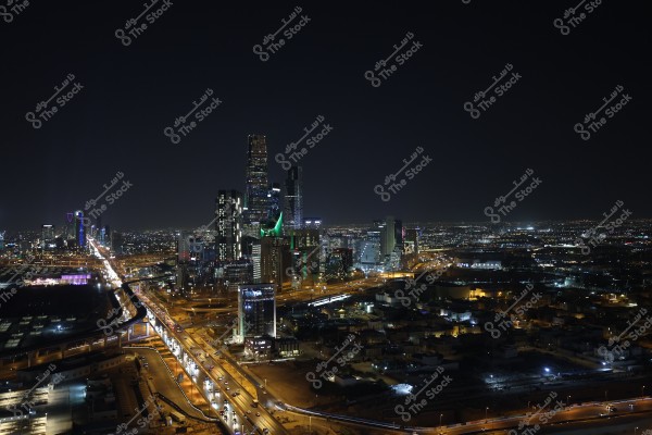 Night view of Riyadh city in Saudi Arabia, showcasing the city skyline adorned with lights. Tall illuminated buildings and the famous tower with its triangular design are visible. The highways on the ground are lit, with cars moving along them.