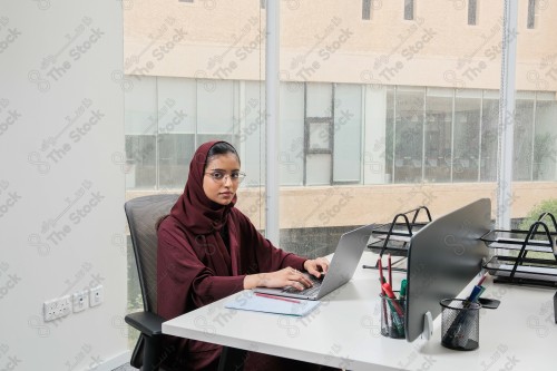 A Saudi woman wearing an abaya works in a glass-fronted office and uses a mobile device to write while taking notes during the day