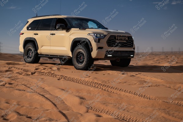 A beige SUV on sand dunes in the desert. The car is shown from the left side with wide front tires and tinted windows. The background includes yellow sand and a set of power towers on the distant horizon under a clear blue sky.