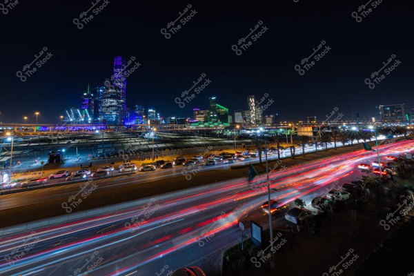 A nighttime image of a city illuminated with brightly colored skyscraper lights, including purple and green hues, with a busy street filled with moving cars, creating light trails due to their speed. The skyline features a cluster of modern buildings, with lights indicating a bustling city life. Cars are lined up on the side of the road under street lighting.