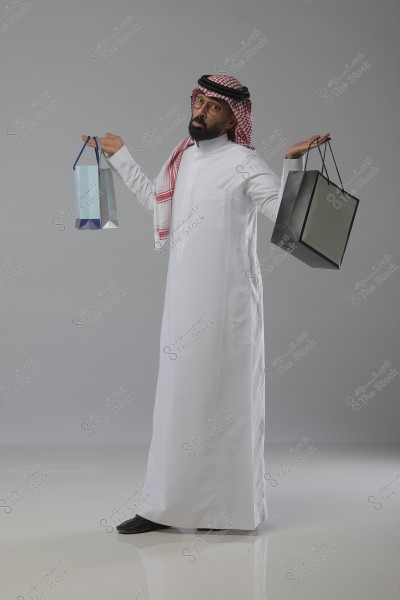 A man wearing traditional Saudi attire, a white thobe and red checkered ghutra, stands in a humorous pose holding shopping bags in his hands. The background is simple and gray.