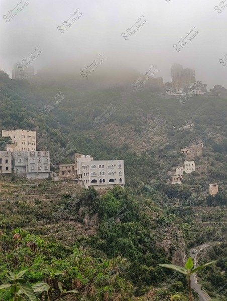 منظر طبيعي لجبل محافظة فيفاء بمنطقة جازان جنوب المملكة العربية السعودية مغطى بالضباب في منطقة جبلية مزروعة بعدة مبانٍ بطوابق متعددة. تظهر المباني ذات الطراز التقليدي بألوان محايدة, محاطة بالخضرة الكثيفة. يتسلل الطريق الضيق الملتف عبر الجبال السفلية, بينما يغطي الضباب القمم العلوية.