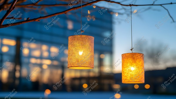Two lanterns hanging from tree branches at dusk. The lanterns emit warm lights, with a backdrop of a lit glass building and blurred lights. The sky in the background is a deep blue, reflecting the evening light.