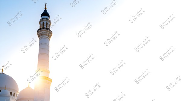 An image showing a mosque minaret with decorative features, accompanied by white domes under a clear blue sky. The sun is rising behind the minaret, creating a bright light effect.