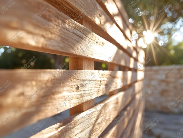 An image of a wooden fence with gaps between the horizontal slats. Sunlight streams through the gaps, creating a beautiful lighting effect and softly illuminating the wooden surface. In the background, there is daylight with blurred trees.