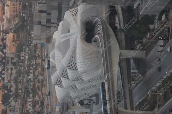 The image shows a modern railway station building in an urban area. The building features an advanced architectural design resembling organic shapes with white panels and curved lines. In the background, multiple buildings, a road with cars, and palm trees are visible.