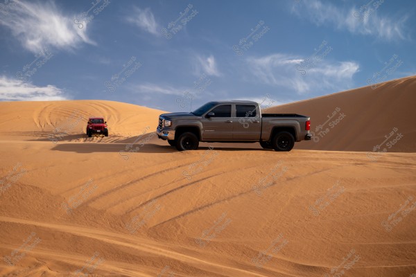 Two off-road vehicles driving on sand dunes in the desert under a clear blue sky with some clouds. The first car is grey and closer to the viewer, while the second red car emerges from the background atop the dunes. The scene depicts an adventurous journey in the desert.