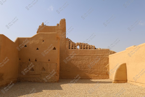 Image of traditional mud-brick walls in an ancient area, showcasing distinctive architectural details with triangular windows and a wall featuring a unique pattern. The walls are beige-colored, likely part of a historical building in Saudi Arabia. The sky is clear blue.