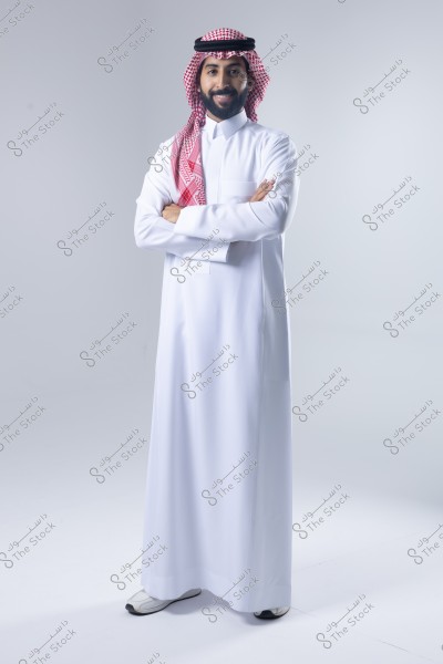 صورة لرجل يقف على خلفية بيضاء مرتديًا ثوبًا أبيض تقليديًا وغترة حمراء مع عقال. الرجل يضع ذراعيه فوق بعضهما البعض ويبدو مبتسمًا. يبدو أن الملابس تشير إلى الزي السعودي التقليدي.