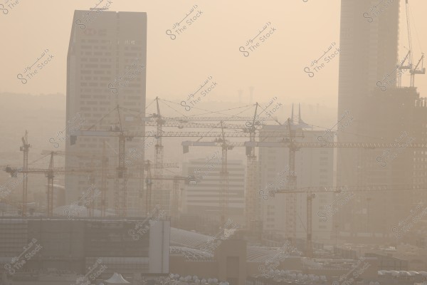 تظهر الصورة مشهدًا لمدينة حديثة تحت الإنشاء مع العديد من الرافعات البرجية المنتشرة في الأفق. تتوسط الصورة أبراج طويلة ومباني شاهقة تحت الإنشاء. السماء مغبرة قليلاً وتظهر ضوء الشمس المتناثر بشكل خافت مما يعطي المشهد مظهرًا ضبابيًا.