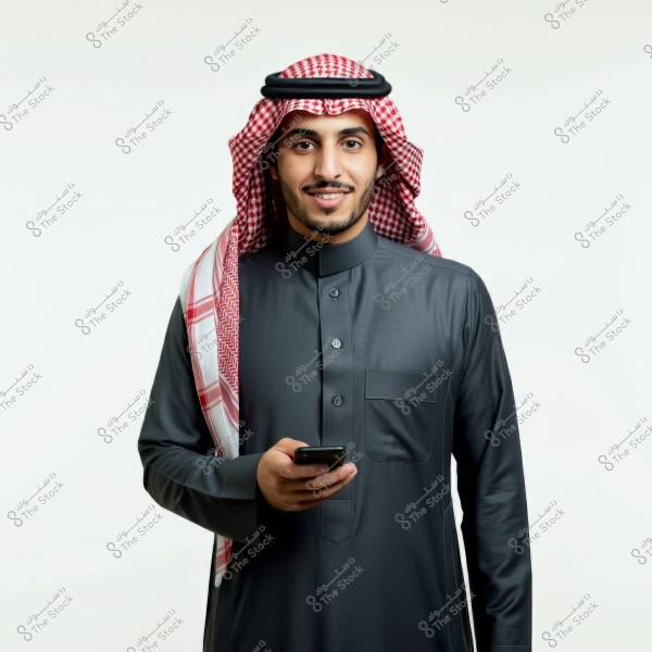 A portrait of a man wearing traditional Saudi attire, including a thobe, ghutra, and agal. The man is smiling and holding a mobile phone in his hand. The white background highlights the details of the traditional clothing.