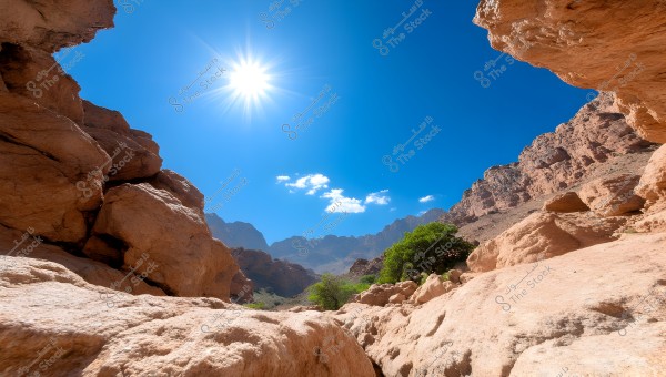 A natural landscape of rocky mountains under a bright blue sky with the sun shining above. Brown rocks and cliffs surround the frame, with limited greenery visible at the bottom. The sky is adorned with a few small white clouds.