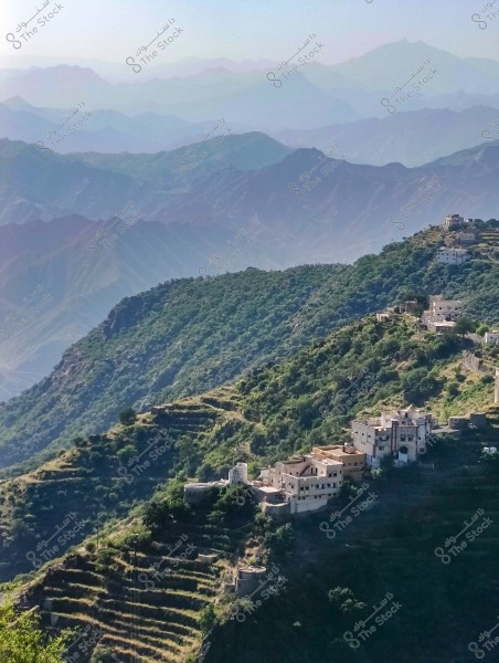 A view of a mountain with a small village in Saudi Arabia, featuring traditional buildings constructed in terrace form. The background shows mountains gradually fading into the distance with a hazy effect.