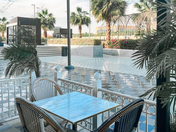 The image shows an outdoor café scene featuring a white table and two woven chairs with metal frames. In the background, a series of palm trees, modern buildings, and lush foliage are visible. The ground is tiled with alternating square patterns. The weather is sunny with a clear sky.