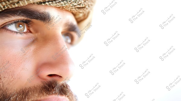A close-up image of a portion of a man\'s face showing one eye and nose with a light beard. He is wearing a headscarf with earthy tones. The background is bright white.