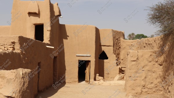 Image of a traditional village built with clay bricks on a sunny day. The picture shows buildings made of clay bricks in a golden-brown color, featuring rectangular and triangular-shaped doors and windows. Trees and plants are visible in the background, suggesting an oasis or desert environment surrounding the site.