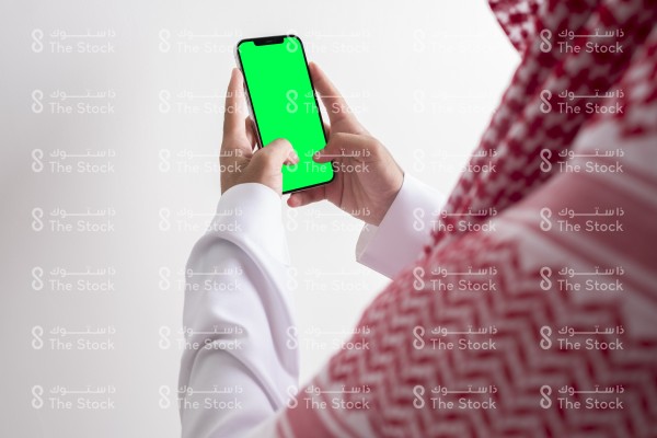 صورة ملتقطة بهاتف من الخلف لرجل سعودي يرتدي الزي السعودي التقليدي يستخدم الجوال وتظهر شاشة الجوال بوضوح