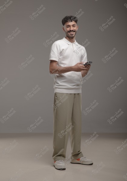 A portrait of a smiling man holding a smartphone. He is wearing a white polo shirt, beige trousers, and sneakers. The simple grey background provides a professional and balanced setting for the image.