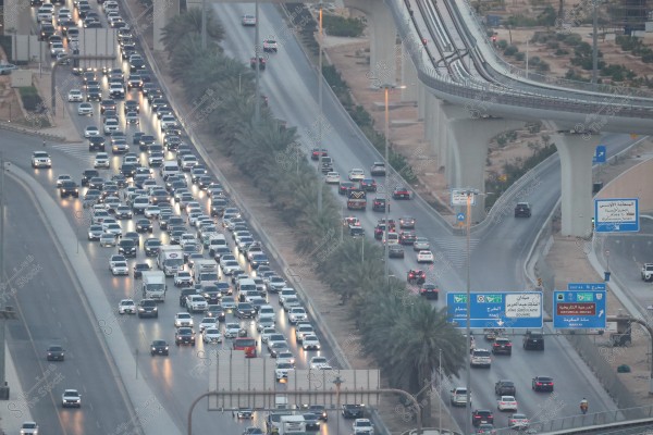 The image shows a traffic jam on a highway in a major city, surrounded by numerous vehicles on both sides, some stationary and others slowly moving. On the left side, there is a road lined with palm trees separating the lanes. An overpass runs across the road, with multiple traffic signs in Arabic and English visible.