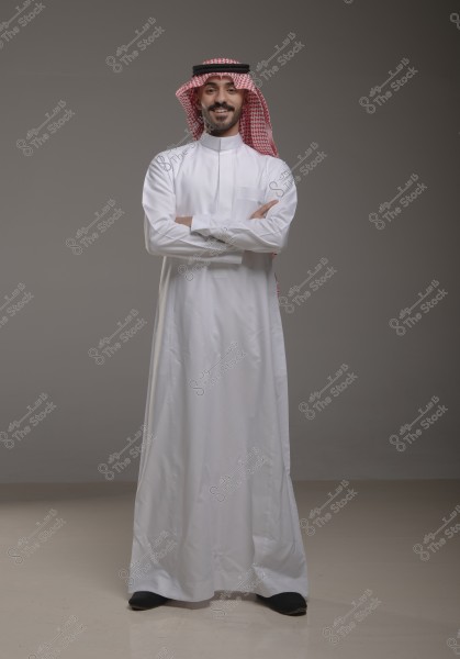 A portrait of a man standing and smiling, wearing a white thobe and a red ghutra with a black agal, suggesting traditional attire from the Gulf region. The man stands upright with his arms crossed, and the background is grey and neutral.