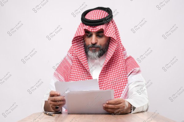 رجل يرتدي الزي الخليجي التقليدي يقرأ أوراقًا على طاولة خشبية.
