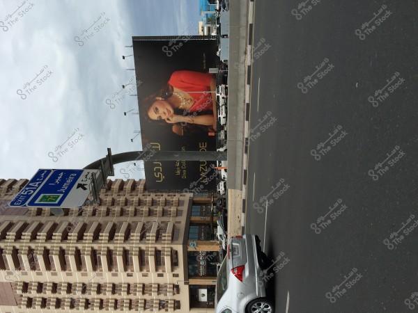 The image shows a view of a city street with a large billboard on a building. The billboard features a woman wearing elegant clothing, gazing to the side. In the foreground, there is a traffic sign indicating the \"Jumeira\" exit. A car is driving on the road, and tall buildings surround the scene.
