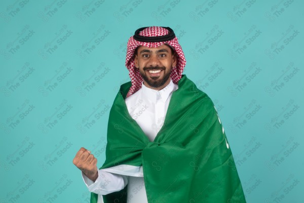 بورتريه رجل سعودي مبتسم يحمل علم السعودية على كتفه احتفالاً باليوم الوطني على خلفية تركاوزية، احتفالات، اليوم الوطني.