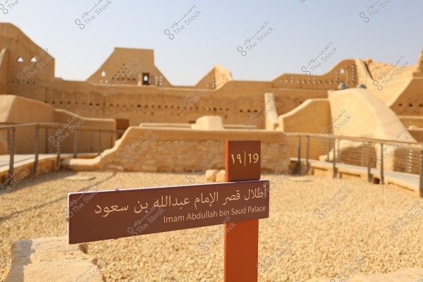Image of a historical site showing the ruins of Imam Abdullah bin Saud Palace. In the foreground, there is a sign with \"Imam Abdullah bin Saud Palace\" written in Arabic and English, along with the number 19,19. The background features traditional mudbrick buildings, adorned with ancient architectural elements. The dominant color is beige, reflecting the heritage architectural styles.