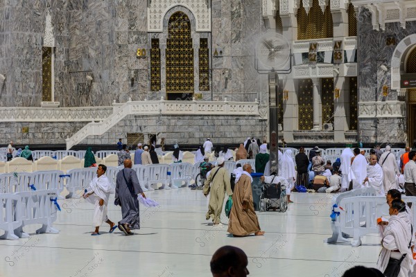 An image depicting a scene in the courtyard of the Masjid al-Haram in Mecca. It shows a group of people wearing Ihram and traditional clothing, some walking and others sitting in the area. The image highlights the distinctive architectural details of the mosque\'s ornate walls and large doors. A white plastic barrier is in the foreground, along with a misting fan and some people in wheelchairs.