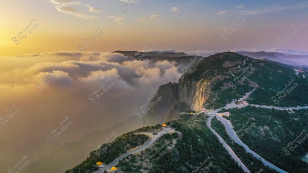 Aerial view of mountains covered in clouds at sunset. A winding road traverses the scene, surrounded by lush greenery, while the sun rises on the horizon, illuminating the sky with golden and orange hues.