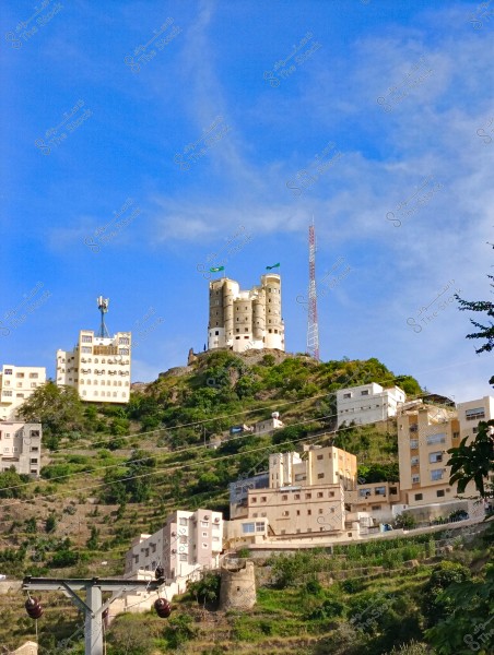 The image shows a fortified castle built atop a hill surrounded by greenery and buildings. The castle has two green flags, and there is a communication tower nearby. Several residential buildings in white and light shades are integrated into the hill below the castle, indicating the mountainous nature of the surrounding area. The sky is blue and clear in the background.