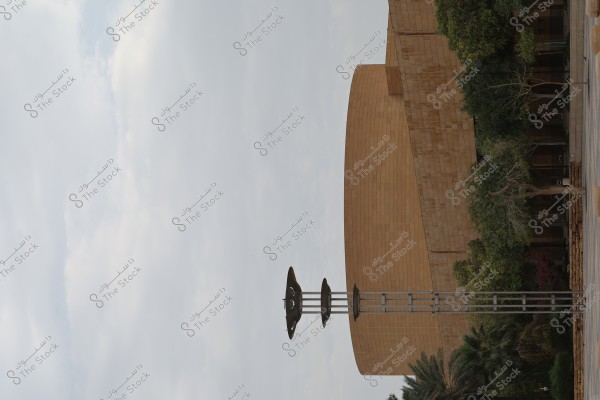 An image showing part of a building with brown brick walls and a curved roof. In front of the building, there is a collection of green trees and ornamental plants. White clouds are visible in the sky.