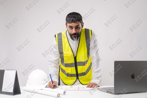 Image of a man wearing a yellow safety vest standing in front of a desk. The man is dressed in a traditional white robe, typical of a Middle Eastern country. On the desk, there is a white helmet, blueprints, an open laptop, and a date planner. The man is holding a pen and writing on the blueprints.
