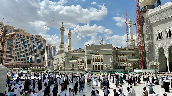 صورة لساحة المسجد الحرام في مكة المكرمة, مليئة بالمعتمرين والمصلين الذين يلبسون ملابس الإحرام. توجد خلفية للمسجد بتصميمه المعماري الفريد والمآذن الشاهقة, ويظهر في الأفق السماء الزرقاء مع بعض السحب البيضاء المنتشرة.

-