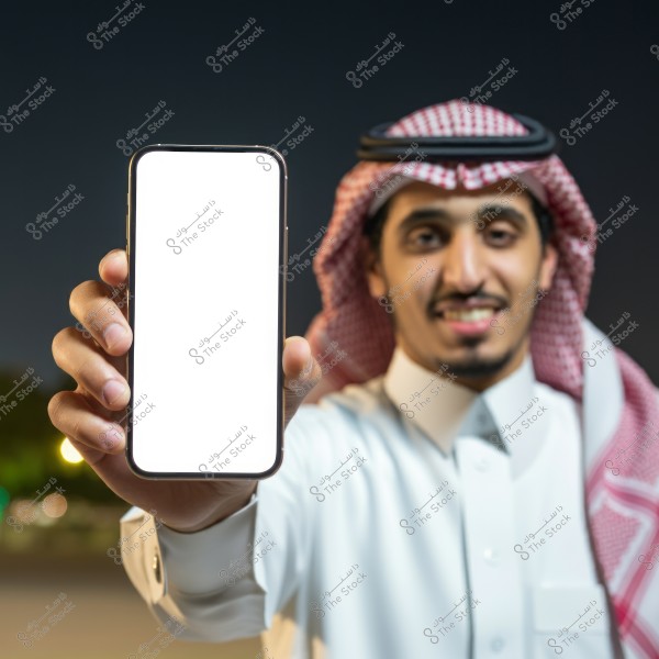 A portrait of a man wearing traditional Saudi attire, including a white thobe and a red and white checkered ghutra with a black agal. He is standing at night and holding a smartphone with a blank white screen towards the camera while smiling.
