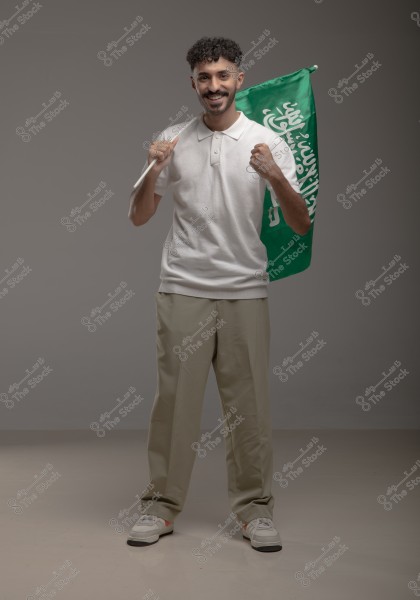 A smiling man stands holding a green flag with white writing. He is wearing a white polo shirt, beige pants, and white sneakers. The flag appears to represent the flag of Saudi Arabia. The background is grey.