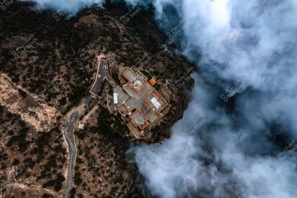 Aerial view of a mountainous area featuring buildings with traditional designs surrounded by trees. Dense fog rises on the right side, adding a mysterious beauty to the scene. A paved road gently winds downward through the trees.
