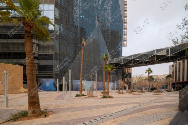 A modern public square featuring palm trees and an open tiled area, surrounded by tall glass buildings. There is a glass overhead walkway connecting the buildings. The atmosphere is calm with a few plants and shrubs.