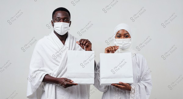 A photo of a man and a woman standing side by side, both wearing white clothing and medical face masks. The man is dressed in a traditional ihram garment, suggesting a Saudi style, while the woman wears a white hijab and an embellished abaya. They are both holding white shopping bags against a white background.