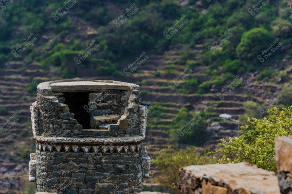 An image of an old partially ruined stone tower in the foreground, featuring a circular design and triangular decorations on the walls. The tower is set amidst a natural mountainous environment covered with greenery and terraced fields in the background, highlighting the traditional character of the area.