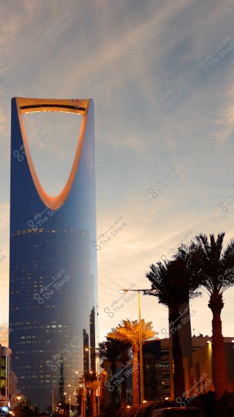 The image shows the Kingdom Tower in Riyadh, Saudi Arabia, during sunset. The tower has a modern architectural design with a large arch at the top, surrounded by palm trees that add to the visual appeal. Warm lights give a tranquil ambiance to the city.
