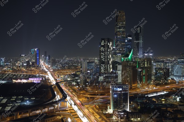 Night view of Riyadh city, showcasing beautifully illuminated skyscrapers and towers. The iconic Kingdom Tower with its blue lighting is visible, surrounded by other buildings lit with colorful neon lights, with busy streets stretching across the city.
