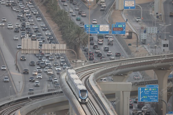 Aerial view of a busy highway in Saudi Arabia, showing numerous cars on the road alongside an elevated metro line with trains in motion. Road signs in both Arabic and English indicate various directions such as \"King Abdulaziz Square,\" \"Dammam,\" and \"Makkah.\" Palm trees are lined along the roadside.